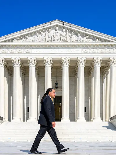 A man with dreadlocks wearing a dark suit and yellow tie walks in front of the Supreme Court Building under a clear blue sky.